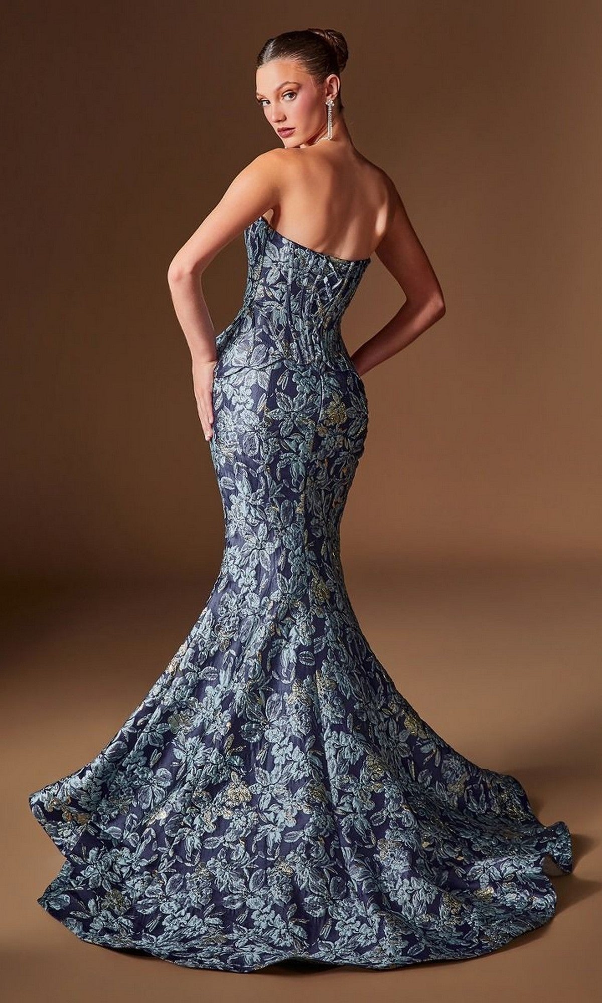 A woman poses against a plain brown backdrop wearing the Ladivine CD655, a strapless, floor-length blue and gold floral jacquard mermaid evening gown featuring a fitted corset bodice.